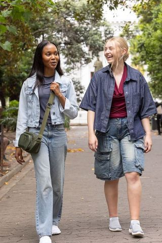 Young women walking and laughing on leafy park path in denim outfits with olive crossbody