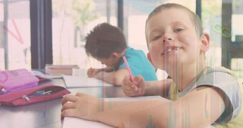 Smiling elementary student writing with pink pencil while classmate studying in bright classroom