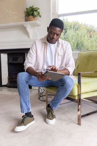 African american man sitting in green armchair using tablet and tapping screen in living room