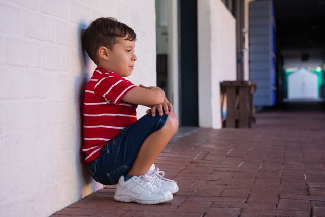 Boy in red shirt and sneakers crouching by a white wall in a school building. Ideal for educational materials, childhood development articles, and school-related advertisements.