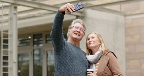Mature couple taking selfie outdoors with coffee near modern urban building