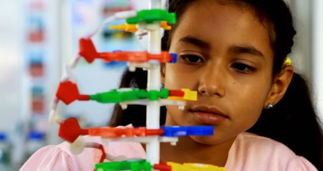 This photo shows a young girl intently studying a DNA molecule model, commonly used in science classes to teach genetics and biotechnology. The colorful model helps simplify complex scientific concepts, making it ideal for educational environments. Use this image for advertisements, articles, and blogs about education, science, children's learning, and educational programs aimed at young minds.