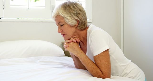 Senior woman with white hair kneeling by bedside in bright room, hands clasped in prayerful contemplation. This image can be used for content related to spirituality, faith, aging, and personal devotion. Ideal for websites, brochures, and articles discussing religious practices, senior living, inner peace, and personal reflection.
