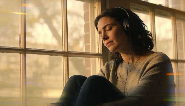 Contemplative woman listening to music on windowsill with headphones in warm sunlight