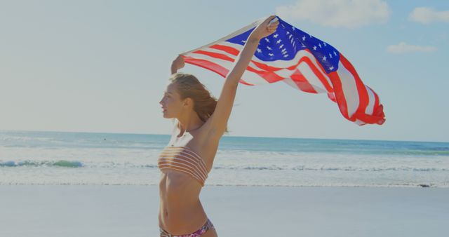 A young Caucasian woman is holding an American flag high as she walks along a sunny beach, with copy space. Her expression of freedom and joy encapsulates a patriotic spirit against the backdrop of the ocean.