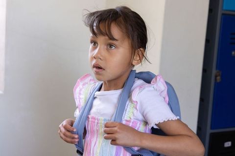 Young girl holding backpack straps looking up with curious expression in school corridor