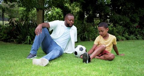 African american father and son sitting on grass sharing soccer ball and bonding outdoors