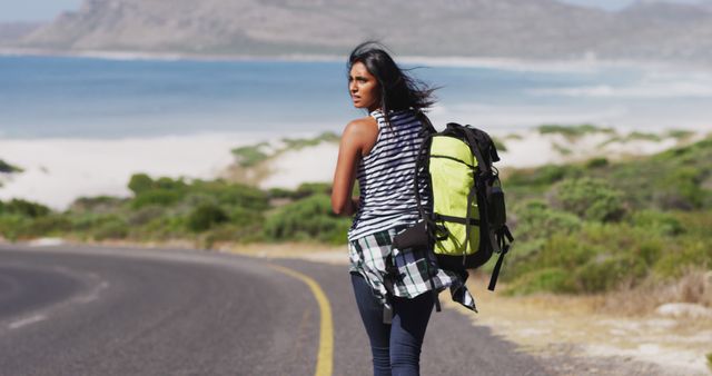 African american woman with backpack trying to hitch a lift while walking on road. road trip travel and adventure concept