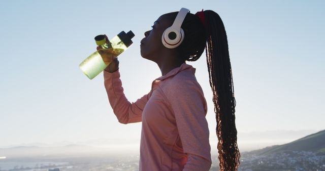 Woman hydrating with water bottle during outdoor workout on a mountaintop. Suitable for promoting fitness, hydration, wellness, and active lifestyles. Ideal for fitness blogs, health magazines, and advertisements about outdoor activities, sports drinks, or workout gear.