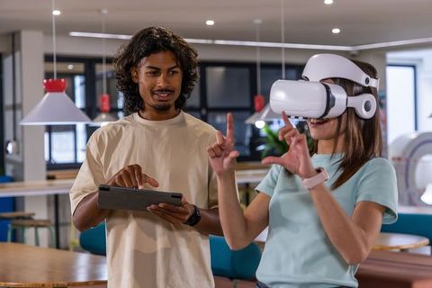 Asian woman testing vr headset while south asian man guiding with tablet in coworking hub