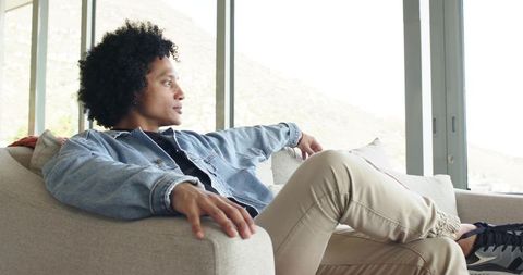 Mid-adult man reclining on light sofa wearing denim jacket, gazing out large windows