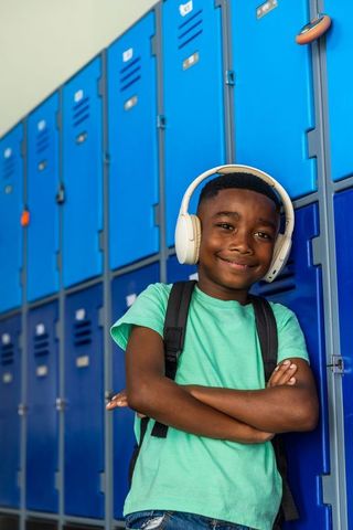 Confident african american schoolboy standing by blue lockers wearing headphones, backpack