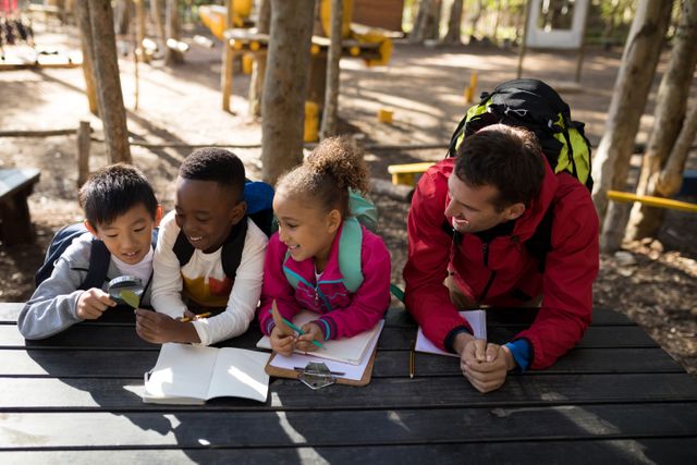This image depicts a teacher guiding a group of children in an educational activity in a park. The kids appear engaged and curious, using tools like a clipboard and magnifying glass to explore nature. This photo can be used in educational content, teaching resources, outdoor learning promotions, and diversity in education themes.