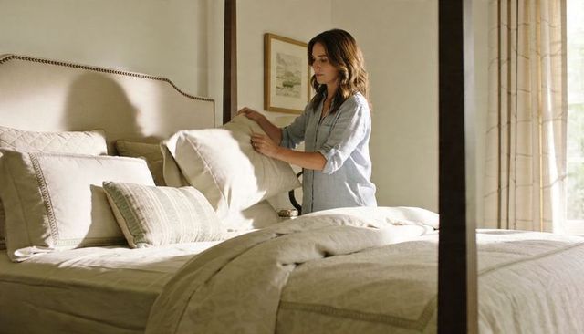Woman making bed in sunlit neutral bedroom arranging pillows for cozy tidy look
