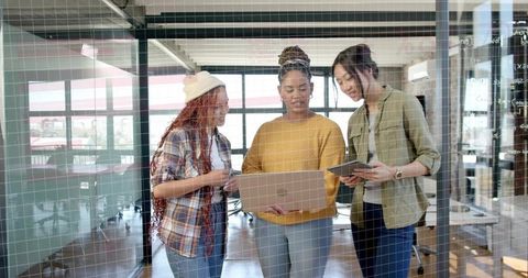 Diverse women collaborating with laptop and tablet behind glass wall in modern office