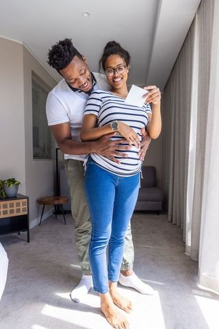 Expecting diverse couple embracing by window in sunlit living room holding ultrasound