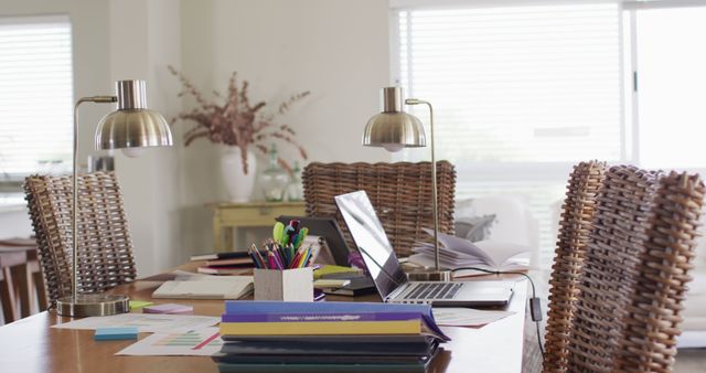 This image depicts a well-organized home office setup with laptops, colorful stationery, and books on a wooden table. The workspace is well-lit by natural light coming from a nearby window, adding a bright and welcoming atmosphere. The office chairs and decorative plants on the table create a cozy ambiance. Ideal for depicting the concept of remote work, productivity environments, or studying from home.