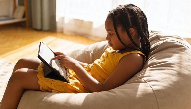Young girl relaxing on beige beanbag tapping tablet wearing yellow dress in sunlit room
