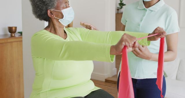 Senior African American woman engaging in exercise with assistance of a physiotherapist at home. Both wearing masks, concentrating on using a resistance band for rehabilitation exercises. Ideal for themes related to senior healthcare, physiotherapy, home workout routines, elderly care, medical treatment, and wellness initiatives.