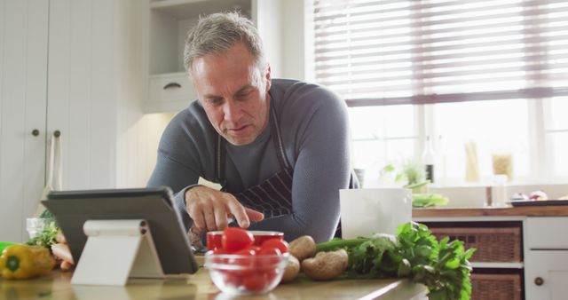Middle-aged man follows a digital recipe on a tablet while preparing a meal in a modern kitchen. Fresh vegetables including tomatoes, mushrooms, and leafy greens are spread on the countertop. Useful for themes related to home cooking, technology in the kitchen, or healthy eating.