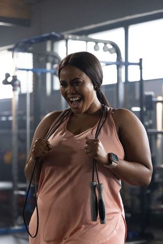 Laughing african american woman holding jump rope in gym wearing pink tank and smartwatch