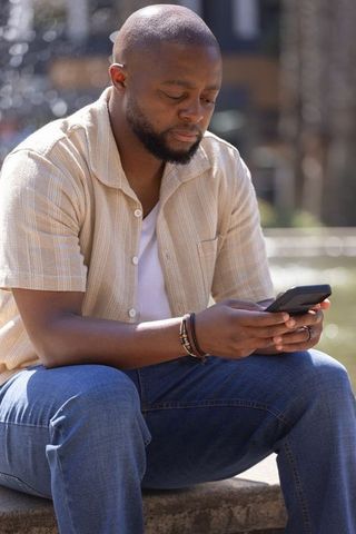 Man sitting at urban fountain holding smartphone, casual beige shirt and bracelet stack