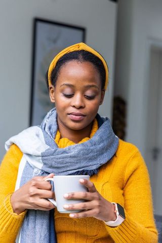 Contemplative african american woman holding mug and looking down in mustard sweater and scarf