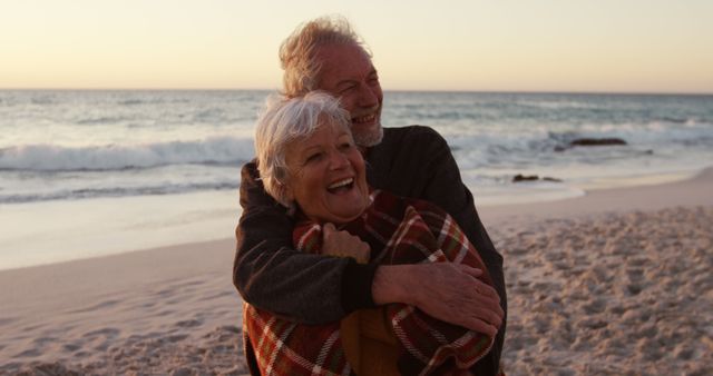 An elderly couple embracing and smiling warmly while standing on a sandy beach at sunset. The ocean waves and the horizon create a serene and romantic backdrop. Perfect for use in advertisements, articles, or promotional materials focused on retirement, love, romance, beach vacations, and healthy aging.
