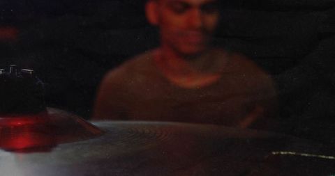 Drummer playing behind cymbal in dim red lighting, moody grainy rehearsal