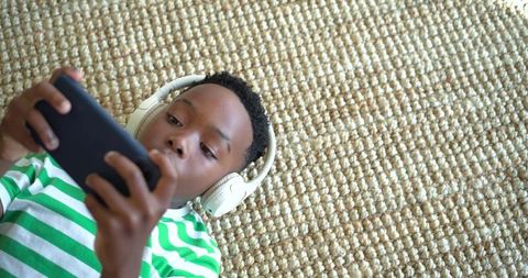 African american boy lying on textured beige rug wearing headphones and using smartphone