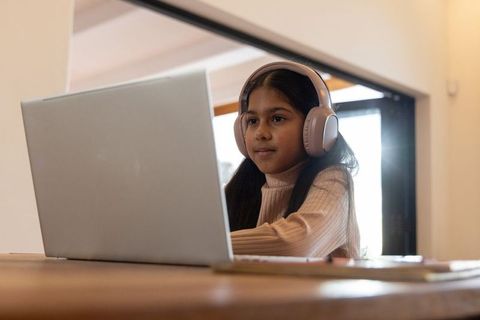 Indian girl learning at laptop with headphones at sunlit wooden desk in calm loft