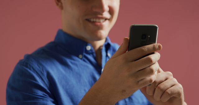 Middle-aged man with casual blue shirt using smartphone against pink background. He is engaged with his phone, likely texting or browsing. This is perfect for illustrating modern communication, technology usage, social media interaction, or lifestyle topics related to connectivity and digital devices.