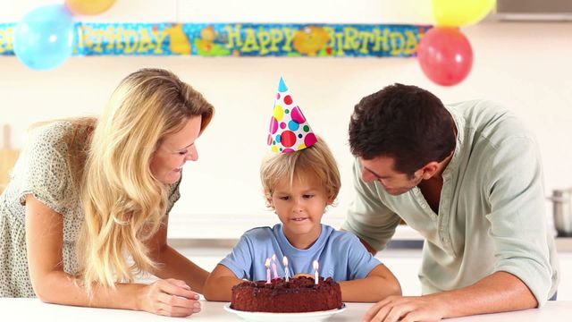 Boy wearing colorful party hat surrounded by parents smiling and blowing out candles on birthday cake at home. Scene perfect for showcasing family togetherness and joyful celebrations. Ideal for social media, blog posts on parenting, or advertising family-friendly events.