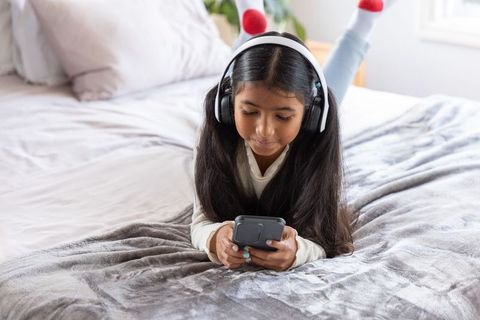 Indian girl lying on bed wearing headphones and holding smartphone, cozy sunlit bedroom