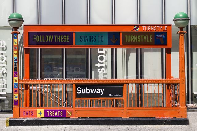 This image showcases a vibrant and eye-catching subway entrance featuring bold orange railings and colorful signage directing commuters. Ideal for use in articles or advertisements focused on urban living, public transportation systems, or city travel guides.