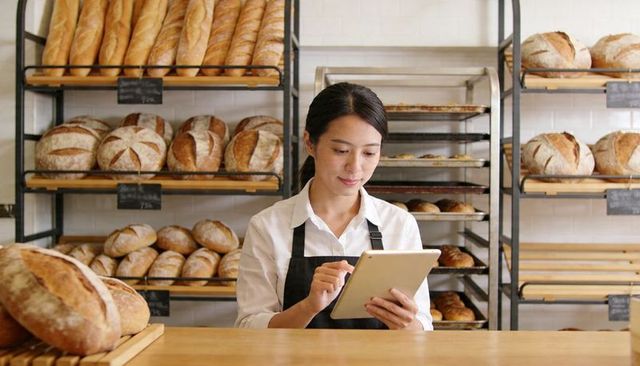 Asian Baker Using Tablet Behind Artisan Bread Shelves, Small Bakery Owner Managing Orders