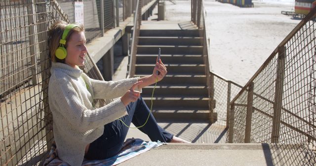 Person sitting near beach engaged in video call wearing headphones and smiling. Ideal for use in advertisements, blog posts, and social media content related to technology, outdoor leisure activities, communication, and youth lifestyle.