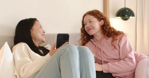 Cozy conversation between Asian and red-haired women on sofa sharing smartphone in minimalist home