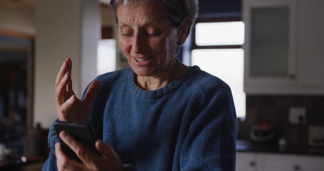 Senior woman in casual clothing using smartphone for video chat while smiling at home. Suitable for concepts like technology use among seniors, staying connected, modern communication, family interaction and elder tech adoption.