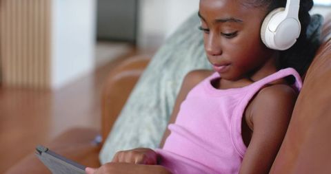 African american girl wearing white headphones using tablet on brown leather couch
