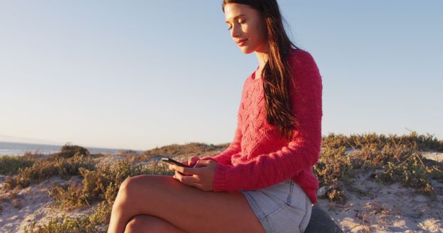 Young woman sitting on a sandy beach at sunset, checking her smartphone. She is wearing a red sweater and denim shorts, enjoying the serene coastal environment. Ideal for use in lifestyle, technology, travel, or relaxation-themed content.