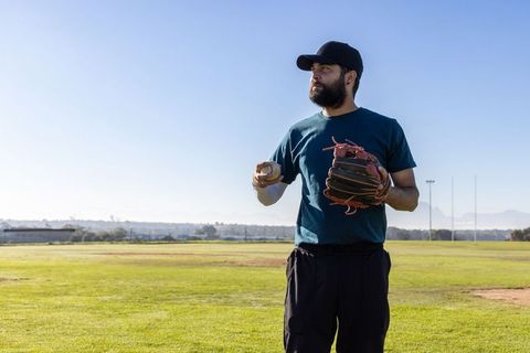 Mid-adult Asian man practicing baseball on sunny field holding ball and glove