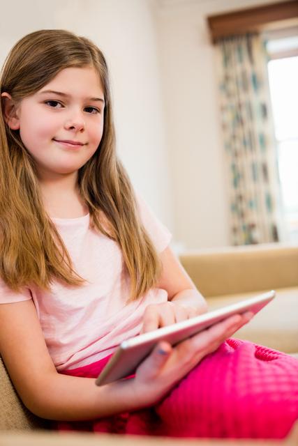 Young girl sitting on a sofa in a living room, using a digital tablet. She is smiling and appears relaxed, suggesting a casual and comfortable environment. This image can be used for themes related to technology, education, modern lifestyle, and children's activities at home.