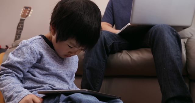Young boy engrossed in using a digital tablet while sitting on floor. In background, parent working on a laptop, suggesting multitasking or shared digital activity. Useful for illustrating family technology usage, remote work scenarios, or modern parenting topics.