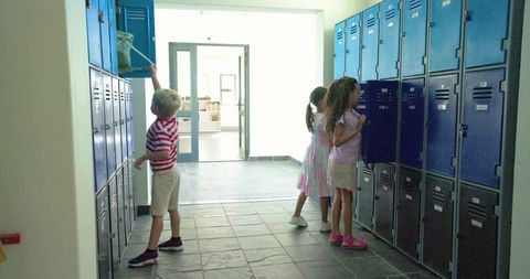 Elementary students reaching into blue lockers using long-handled net in bright hallway