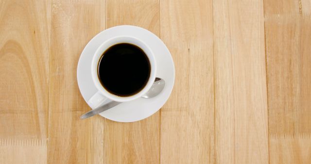 This image showcases an overhead view of a cup of black coffee placed on a white plate with a spoon on a light wooden table. Ideal for use in coffee shop promotions, websites, blogs about morning routines, and design materials focused on simplicity and minimalism.