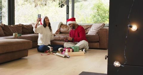 Diverse couple wrapping gifts and video calling in cozy living room wearing Santa hats