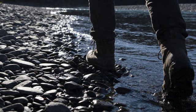 Hiker walking boots splashing through pebble riverbank with glistening water