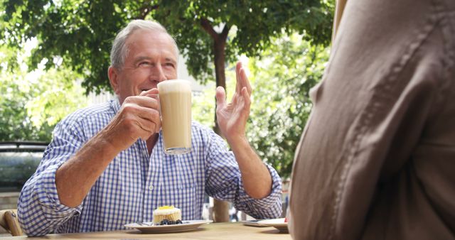 The senior man is seen enjoying a coffee and desserts while engaging in a friendly conversation at an outdoor café. Suitable for use in themes related to relaxed lifestyles, social interactions, senior well-being, family gatherings, leisure activities, and outdoor events.