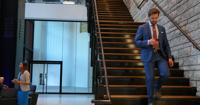 A Caucasian businessman in a suit is focused on his phone while descending stairs, with copy space. A Caucasian businesswoman in the background appears engaged in her own mobile device, highlighting the common use of technology in modern corporate settings.